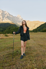 Fototapeta premium A confident outdoor enthusiast stands in a grassy alpine meadow with a trekking pole, wearing a jacket, shorts, and boots. Sunlit mountains rise behind, creating a vivid adventure scene.