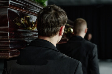 Caucasian young adult men carrying wooden coffin during funeral ceremony, walking in line with focus on back of heads, formal attire visible, somber indoor setting