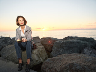 A confident professional woman sits on rugged rocks at sunset, texting on a smartphone in smart casual attire, creating a calm, focused mood by the sea.