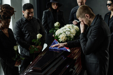 Group of middle aged and young adult multiethnic men and women mourning around coffin, Caucasian man wiping tears while touching folded flag, people holding roses