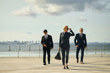 Caucasian middle aged businesswoman in business suit and sunglasses walking confidently outdoors with two male colleagues in suits following behind near waterfront urban background