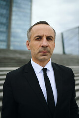 Portrait of middle aged Caucasian businessman, looking into camera, standing outdoors in front of corporate headquarters on office building steps, wearing formal suit and tie with serious expression