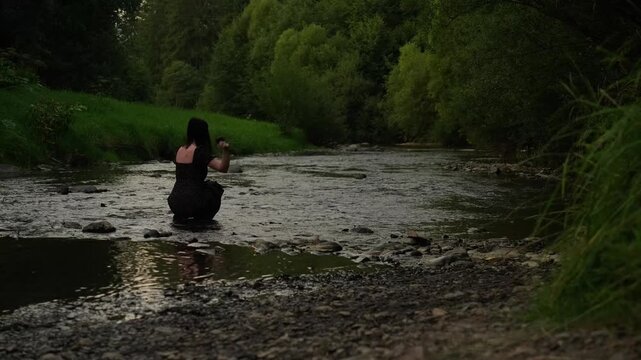 A woman splashes water with her hand and throws stones in a mountain river in the evening