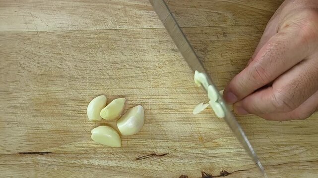 Man hands chopping raw garlic on a scratched wooden cutting board with a kitchen knife. Top view