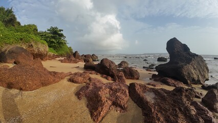 Scenic coastal landscape with large rocky formations and tropical greenery along a sandy beach under a bright cloudy sky