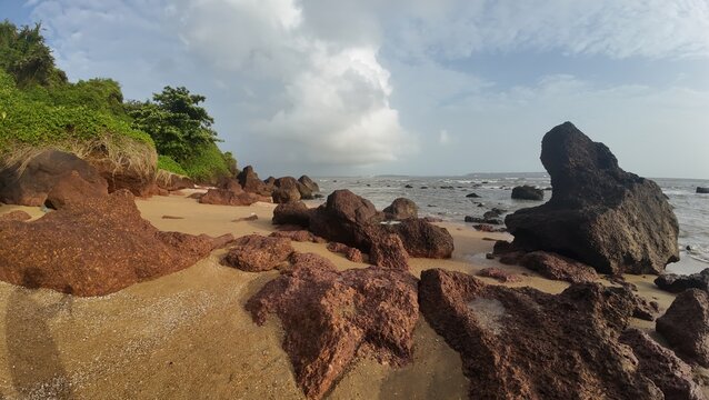 Scenic coastal landscape with large rocky formations and tropical greenery along a sandy beach under a bright cloudy sky