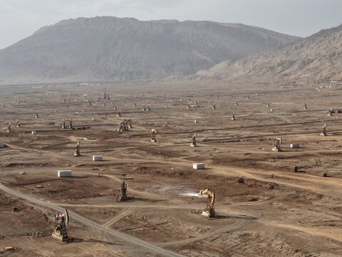 Aerial view of a desolate, dusty landscape dotted with oil derricks stretching towards hazy mountains, a scene of industrial activity against a backdrop of raw, arid beauty, Turpan, Xinjiang, China.