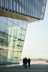 Fototapeta premium Three middle aged professionals walking side by side outdoors near modern glass building, carrying briefcase, business district skyline visible in background, engaging in conversation