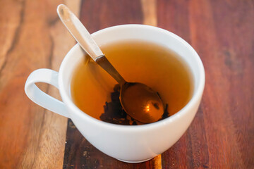 A white ceramic cup of amber tea with a metal spoon rests on a richly grained wooden surface, evoking warmth and simplicity.
