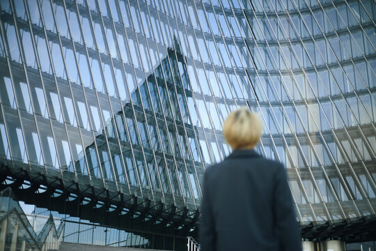 Caucasian young woman in formal suit standing outdoors facing modern glass office building, observing architectural reflections on curved facade, career, motivation, ambition and achievement concept