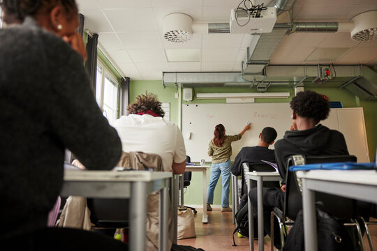 Low angle rear view of students sitting at desk while female teacher erasing whiteboard in classroom