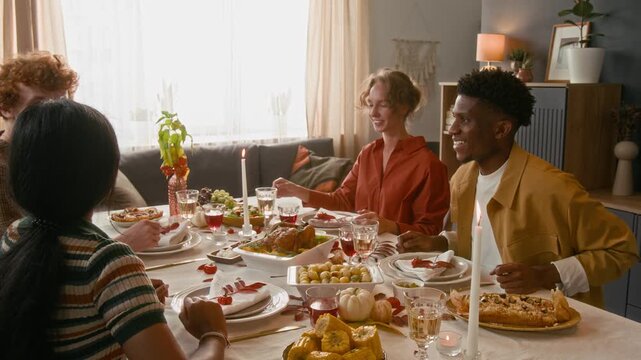 Group of multiethnic young friends finishing grateful prayer and getting down to holiday meal while sitting at beautifully set dining table in living room