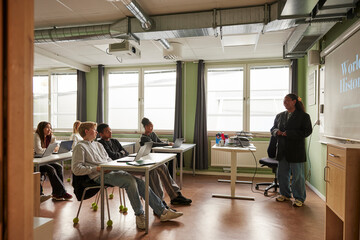 Female teacher teaching about world history on projection screen to junior high students sitting with laptops in classro