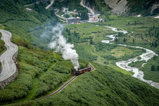 Aerial view of a vintage steam train chugging through verdant hills alongside a winding road, with the village of Obergoms nestled in the valley, Obergoms, Valais, Switzerland.
