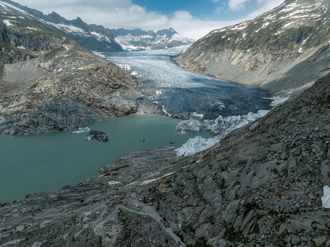 Aerial view of the Rh&Atilde;&acute;ne Glacier cascading into a milky lake, framed by rugged, rocky mountains and snow-capped peaks under a vast sky, Obergoms, Valais, Switzerland.
