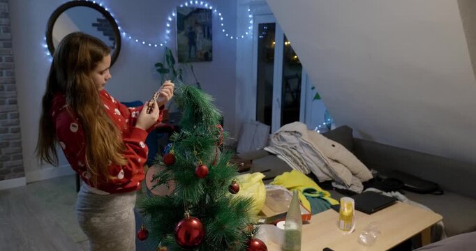 Daughter decorating the Christmas tree, holding a star in her hand, at home at night. Wide shot.