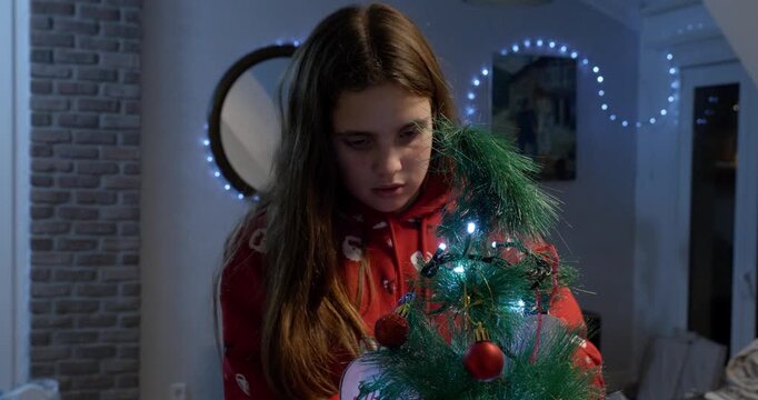 Close-up of a girl wearing red Christmas clothes, putting decorations on the Christmas tree. The focus shifts from her face to the tree.