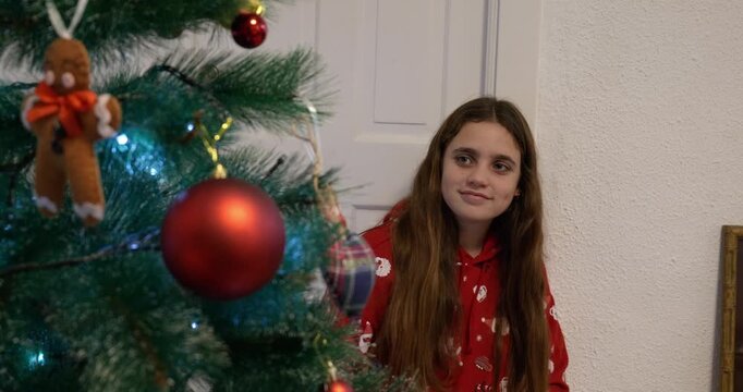 A girl dressed in Christmas clothes turns on the Christmas tree lights at home and looks at it with a satisfied expression. Tree in the foreground.
