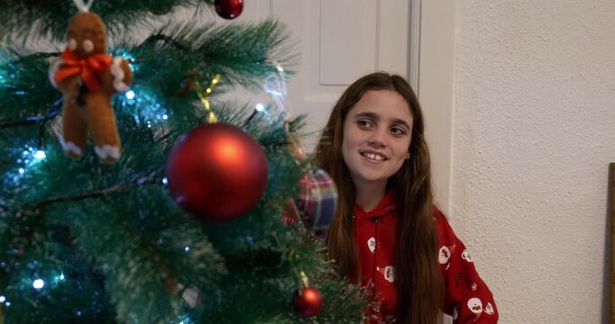 Girl in Christmas clothes looks at the Christmas tree at home and gazes at it with a satisfied expression. Tree in the foreground.  