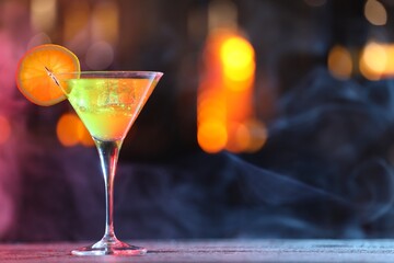 Glass of alcoholic cocktail with garnish on table against blurred background with smoke, space for text. Bokeh effect