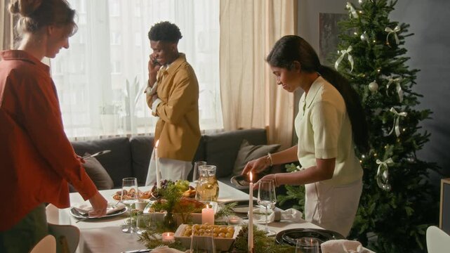 Two multiethnic girls and young man setting festive table with dinnerware while getting ready for Christmas meal, Black guy speaking on phone in background