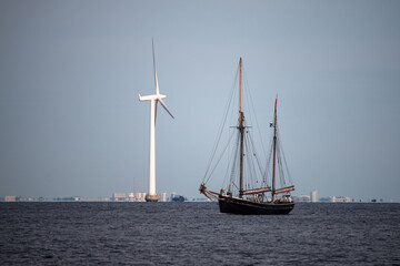 Historic wooden sailboat sails past a towering offshore wind turbine in calm waters, symbolizing the evolution of marine energy.