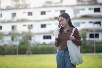 Fototapeta premium Photo of an Asian woman standing holding a paper cup and carrying an eco-friendly bag in a park.