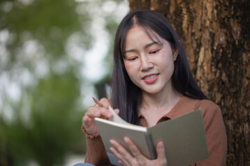 A young Asian woman writes or reads a book under a tree, happily using eco-friendly materials