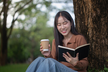 A young Asian woman sits against a tree happily reading a book with a paper coffee cup in her hand.
