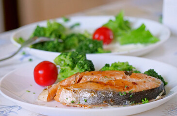 A close-up of a delicious and healthy meal on a white plate. The focus is sharp on the foreground, showcasing the perfectly baked salmon steak, while the broccoli and tomato are slightly softer