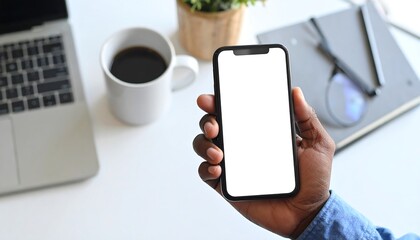 A hand holding a smartphone with a blank white screen on a desk surrounded by work essentials