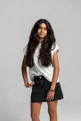 Trendy teenage girl posing with hands in pockets, casual white t-shirt and black denim skirt, cool urban studio portrait.