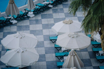 Top view of a poolside area with white parasols, turquoise sun loungers, tiled flooring, and palm tree leaves, creating a relaxing summer resort vibe.