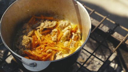 Meat with carrots and ground black pepper is boiled in a pot in nature, the pan is steaming and bubbling with protein broth, tourist recreation, food on the fire, a happy family dinner.