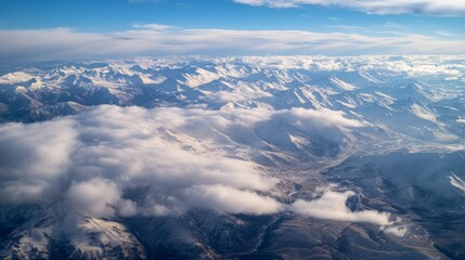 Fototapeta premium Aerial view of majestic snow-capped mountains under a bright blue sky, with fluffy clouds hovering over the rugged terrain, showcasing the beauty of nature and vast landscapes
