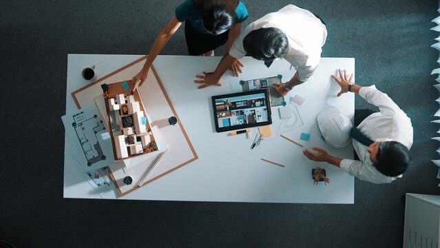 Top down view of architect engineer team discussing and point at tablet display construction at table with blueprint and house model at meeting. Diverse designer planning design house. Alimentation.