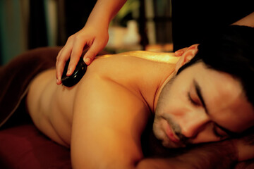 Obraz premium Hot stone massage at spa salon in luxury resort with warm candle light, blissful man customer enjoying spa basalt stone massage glide over body with soothing warmth. Quiescent