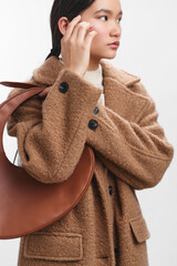 Asian woman wearing a brown wool coat and a curved leather bag stands in a clean studio, adjusting her hair and looking aside with calm, poised confidence.