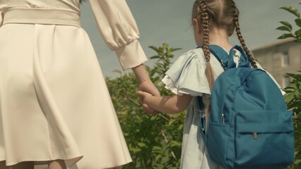 mother leads a small child by the hand to the first grade of elementary school, school bag on shoulders of a girl, happy family, rush to lesson in schoolyard, kid with backpack will get an education.