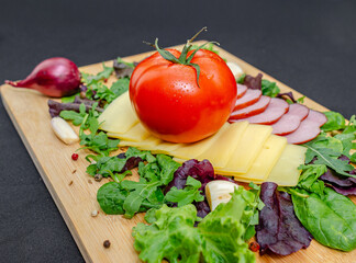 Board with sliced cheese, ham, served with vegetables and herbs on black. Healthy protein menu.