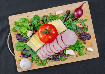 Board with sliced cheese, ham, served with vegetables and herbs on black. Healthy protein menu.