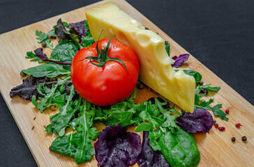 Red tomato and large piece of cheese with greens on wooden board on black. Healthy eating concept.