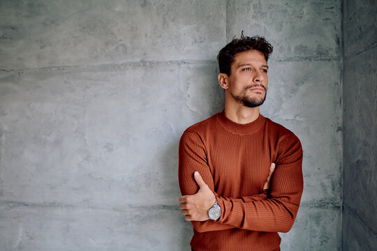 Latino man in an orange ribbed sweater with crossed arms, looking away thoughtfully against a concrete wall