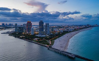 Aerial night view of Miami coastline. Twilight skyline above the Miami oceanfront. Miami Beach waterfront glowing after sunset. South Point lifeguard tower at dusk. Famous Miami Beach from above.