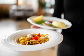 Restaurant-style serving of seafood pasta and grilled meat with sides, held by diner. Gourmet presentation, shallow depth of field, cozy dining ambiance