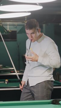 Close-up of a player in white shirt with calm expression, holding cue stick and glass of lemon drink while pacing around pool hall. Overhead lights cast a stylish ambiance over green pool tables