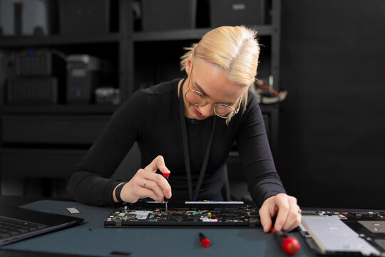 Female IT technician in her 30s repairing a laptop motherboard with screwdriver in a tech lab