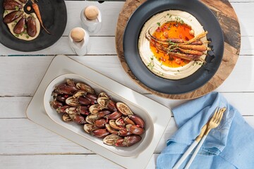A delicious spread of middle eastern inspired dishes is arranged on a white wood table. Hummus is served with roasted eggplants, carrots, and pomegranate seeds