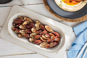 Grilled baby eggplants are presented in an oblong dish with pomegranate seeds sprinkled on top, sitting on a white wooden table with hummus and a blue napkin nearby