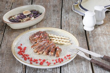A plate holds sliced duck breast adorned with pomegranate seeds, accompanied by grilled baby corn. A dish of cooked eggplant and white salt and pepper shakers complete the rustic scene